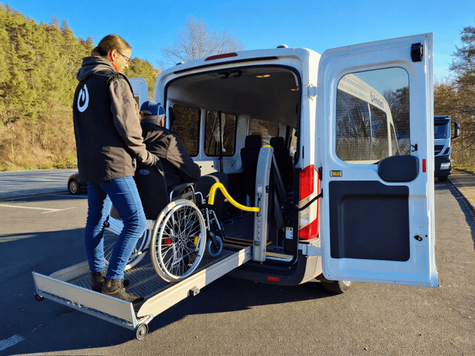 Eine Mitarbeiterin begleitet einen Fahrgast im Rollstuhl über die Rampe in den Kleinbus.